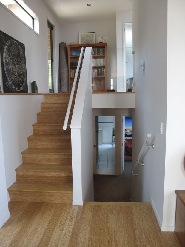 Bright modern interior view of a split-level stairway and landing. Warm timber stairs and flooring contrast with white walls and a sculptural white stair balustrade, with a timber bookshelf and artwork on the upper landing. A lower hallway and tiled room are visible beyond the stair opening.