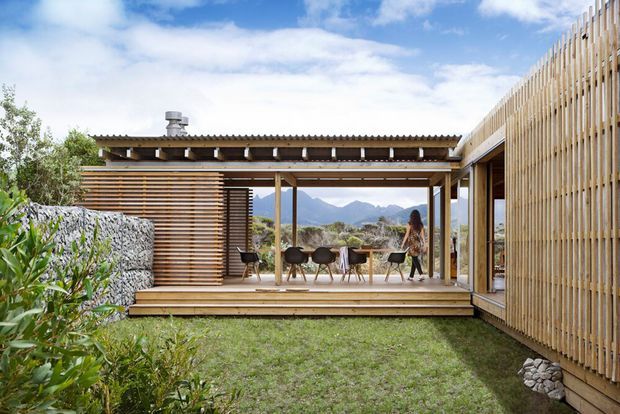 A contemporary timber-clad residential courtyard with an open-air dining terrace beneath a covered pergola. Vertical and horizontal timber screens frame mountain views, with a small lawn, planted garden beds, stone landscaping, and an adjoining glazed volume.