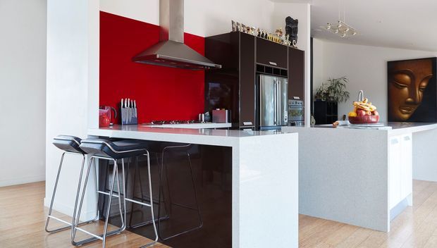 Contemporary open-plan kitchen with a dark brown island and tall cabinetry, white stone-look benchtops, a vivid red splashback, stainless steel rangehood and appliances, and a casual breakfast bar with modern stools. The adjoining space includes a white peninsula, decorative artwork, and timber flooring.