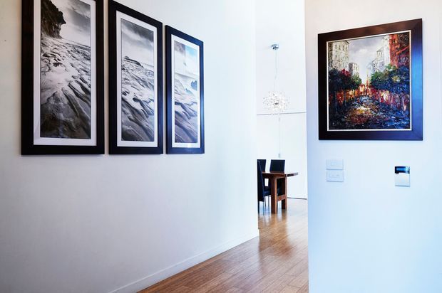 Bright residential hallway with white walls, timber flooring, and a curated arrangement of framed artwork. The hallway opens toward a dining area with a wooden table and chairs, while recessed lighting and wall controls contribute to a clean contemporary interior.