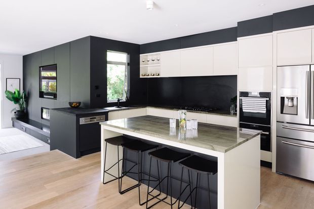 A spacious contemporary kitchen with a large central island and four black bar stools. The room features matte black full-height cabinetry and splashback, white upper and lower cabinetry, integrated appliances, a stainless-steel refrigerator, and a pale stone island benchtop. Light timber flooring and abundant natural light create a clean, refined interior.