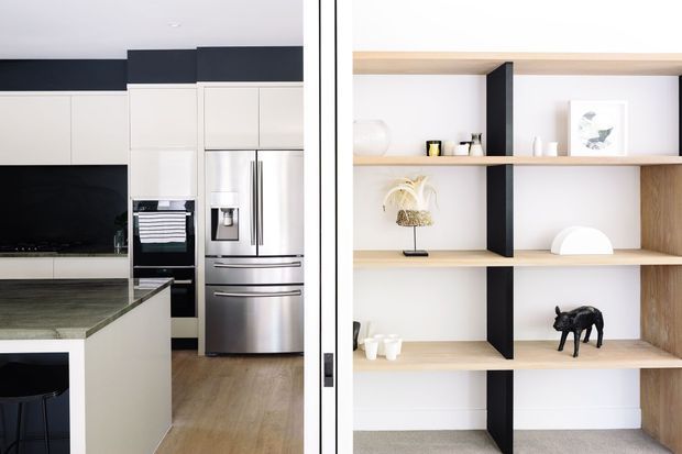 A bright contemporary kitchen and adjoining open shelving display, viewed through a glazed or framed opening. The kitchen features white flat-panel cabinetry, black accents, stainless-steel appliances, a dark stone island benchtop and timber flooring. The adjacent built-in shelving combines light timber shelves with black vertical supports and minimal decorative objects.