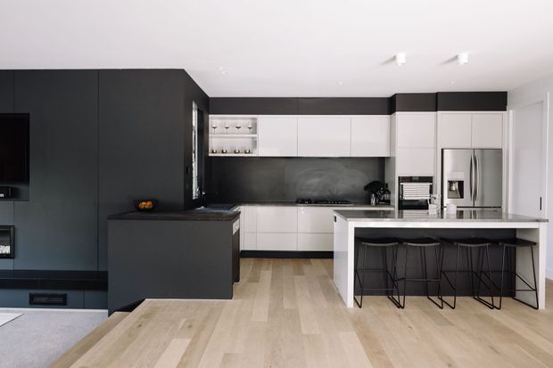 Modern open-plan kitchen with a large island, integrated dark cabinetry, white upper and lower cabinets, stainless-steel appliances, dark splashback, and pale timber flooring. Three black bar stools sit beneath the island benchtop.