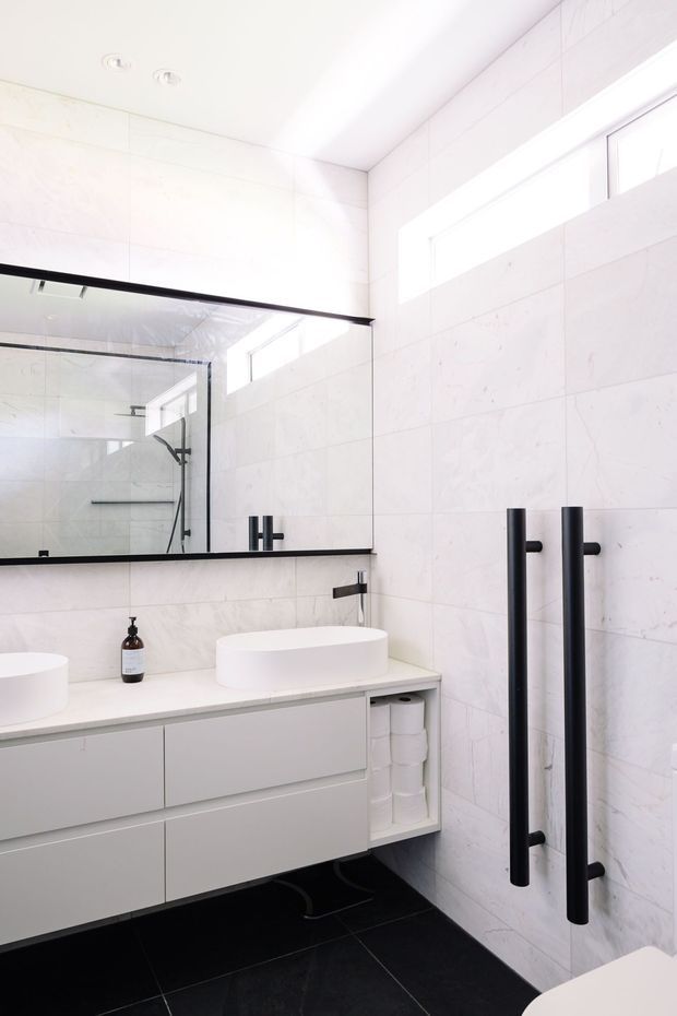 Bright contemporary bathroom with white marble-look wall tiles, a floating white vanity with integrated storage, twin white vessel basins, matte black tapware and tall black heated towel rails. A large frameless mirror reflects the glass shower enclosure, while narrow clerestory windows provide natural light. The floor is finished in dark charcoal tile.