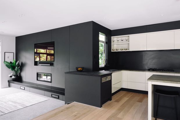 Open-plan contemporary kitchen and living area featuring matte black full-height cabinetry and media joinery, a white kitchen with dark splashback, timber flooring, and a compact island or peninsula with seating. Integrated fireplace and television cabinetry form a dark feature wall, while a large rug and indoor plant soften the space.