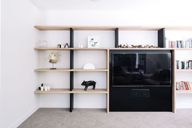Bright, minimalist living room media wall featuring open light-wood shelving with dark vertical supports, a black built-in television and speaker cabinet, books, and curated decorative objects. The room has white walls and soft grey carpet.