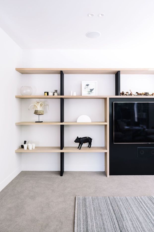 Bright contemporary living room with a built-in open shelving wall in pale timber and black vertical supports. The shelving displays decorative objects and artwork, beside a large black television unit. The room has white walls, recessed ceiling lights, light grey carpet, and a partially visible grey striped rug.