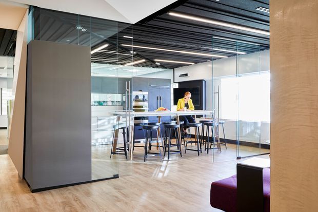 Bright contemporary commercial office interior with glazed meeting rooms, a communal high table and stools, exposed dark linear ceiling services, pale timber-look flooring, and colourful lounge seating. The space has a polished, open-plan workplace character with extensive internal glass partitions.
