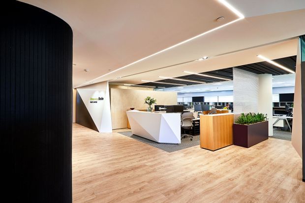 A contemporary commercial office reception area with a sculptural white reception desk, wood-look flooring, warm timber cabinetry, planter boxes, black ceiling slats, integrated linear lighting, and open-plan workstations visible beyond. The space has a polished, welcoming corporate fit-out with strong geometric forms and contrasting light and dark finishes.