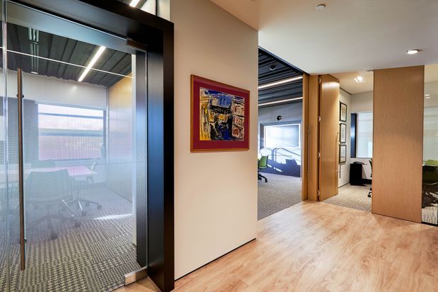 A contemporary commercial office interior viewed from a circulation area. The space features light timber-look flooring, warm neutral walls, black-framed glazed office partitions, a carpeted meeting room visible through glazing, timber veneer doors and panels, and an exposed dark ceiling with linear lighting. Artwork provides a small colourful accent.