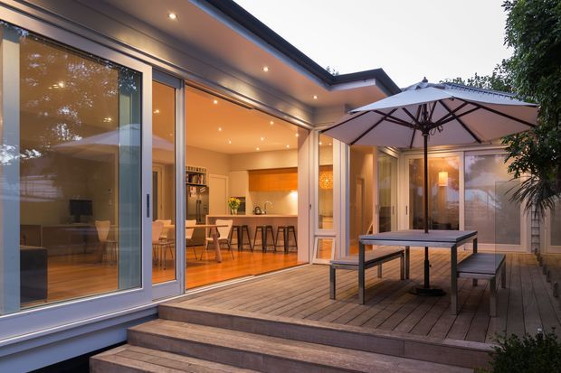 A contemporary residential entertaining area viewed from a timber deck, with extensive sliding glass doors opening into a warmly lit kitchen and dining space. The kitchen features a long island with bar seating, white cabinetry, and a timber-toned feature wall. A covered outdoor dining setting with a large umbrella occupies the foreground, while the surrounding glazed walls and low-pitched roof create a strong indoor-outdoor connection.