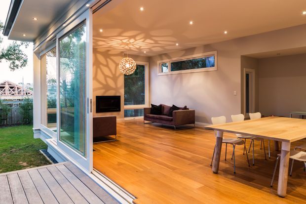 Open-plan contemporary living and dining area extending onto a timber deck through large glazed sliding doors. The room features warm timber flooring, pale walls, recessed ceiling lights, a compact fireplace or television wall, a low dark sofa, and a timber dining table with light chairs. A sculptural pendant light casts an organic patterned shadow across the wall and ceiling. The space opens to a small landscaped yard and appears bright, warm, and connected to the outdoors.