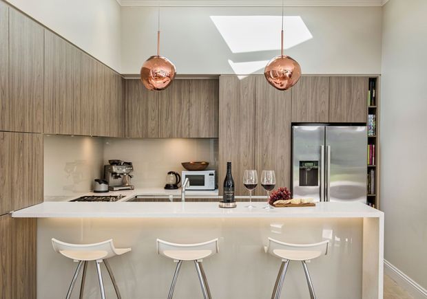 Bright contemporary kitchen with a large white island and seating for three, extensive wood-grain cabinetry, integrated stainless-steel appliances, a pale splashback, and copper pendant lights. A skylight provides strong natural illumination, while classic wall moulding adds a subtle traditional detail.