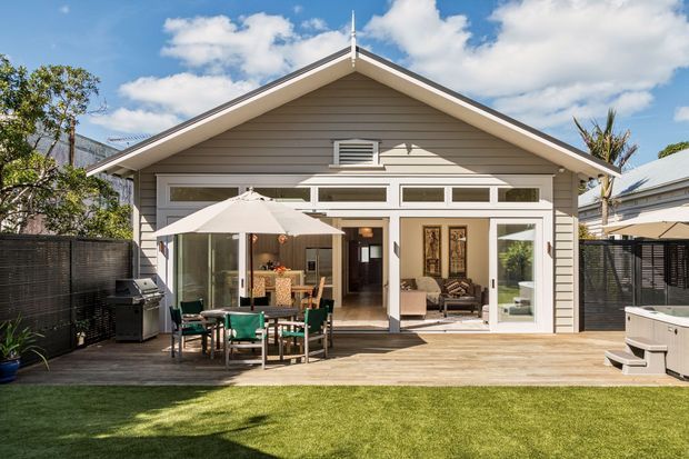 A bright rear exterior view of a single-storey gable-roof home opening onto a timber deck and lawn. The facade features horizontal weatherboard cladding, white-framed glazing and French doors, with an outdoor dining setting, barbecue, umbrella, and spa pool on the deck.