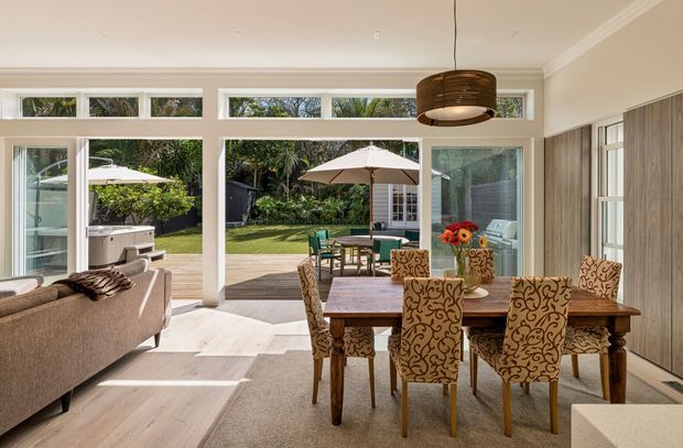 Bright open-plan dining and living area with a large timber dining table, patterned upholstered dining chairs, a neutral sofa, and extensive floor-to-ceiling glazing opening onto a landscaped patio and lawn. The room features light timber flooring, pale walls with simple cornice detailing, a woven pendant light, and built-in timber cabinetry along the right wall.