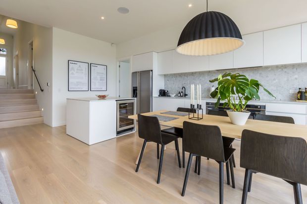 Bright, open-plan contemporary kitchen and dining area with white flat-panel cabinetry, a pale stone-look splashback, light timber flooring, and a large timber dining table surrounded by dark upholstered chairs. A sculptural black pendant light hangs over the dining table, while a central island, integrated appliances, staircase, and framed wall art complete the space.