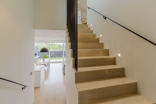 Bright contemporary stairway with broad light timber treads and risers, white walls, slim black handrails, and integrated square wall lighting. The stairwell opens toward a modern dining and living area with large glazing in the background.