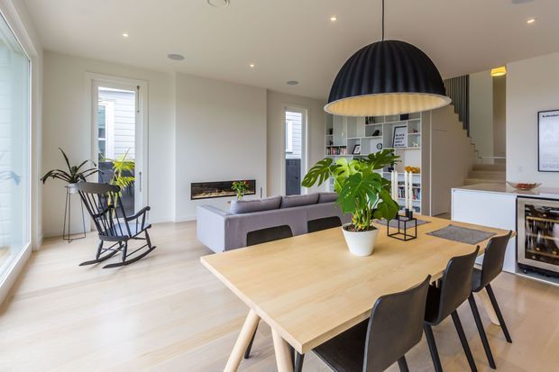 Bright, open-plan contemporary living and dining area with a large light-wood dining table, dark upholstered dining chairs, grey sofa, indoor plants, and a prominent black ribbed pendant light. Built-in shelving and cabinetry sit beneath a staircase, with a compact beverage or wine refrigerator visible at right. Pale timber flooring, white walls, recessed lighting, and large windows create an airy modern interior.