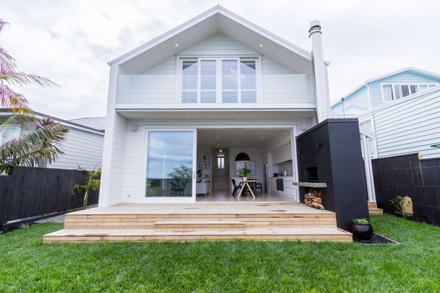 Front exterior view of a compact two-storey coastal-style home with a crisp white gabled facade, horizontal weatherboard cladding, upper-level louvred glazing, and a broad timber deck. The open ground-floor living area connects to the deck, with a prominent black outdoor fireplace and chimney on the right. A fenced lawn and neighbouring weatherboard homes frame the scene.