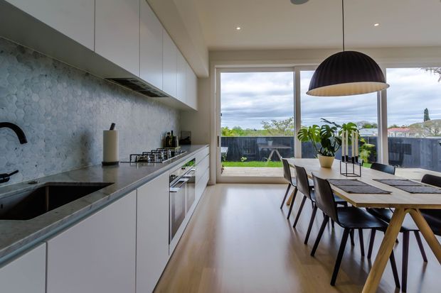 Contemporary open-plan kitchen and dining area with white handleless cabinetry, a pale hexagonal tiled splashback, dark stone benchtops, and warm timber flooring. A large dining table sits beneath a black dome pendant light, while full-height glazed doors open to a fenced garden.