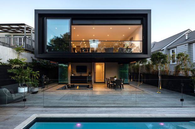 Contemporary two-storey residential rear facade with a dramatic cantilevered upper volume framed in dark cladding and expansive glazing. The lower level opens onto a timber deck and pool terrace, with outdoor dining, built-in seating, a linear fire feature, landscaping, and frameless glass pool fencing. Warm interior lighting contrasts with the dark exterior envelope and dusk setting. Neighbouring residential buildings are visible on either side.