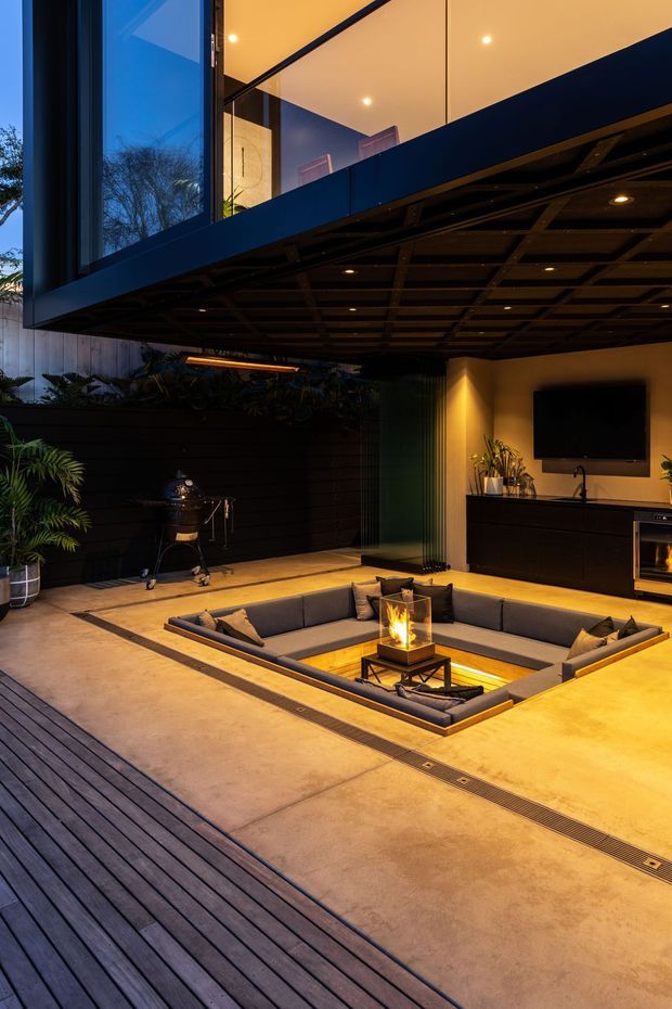 A sophisticated covered outdoor entertainment area at dusk, featuring a sunken U-shaped lounge with a central fire feature, a wall-mounted television, dark built-in cabinetry with a sink and barbecue, timber decking, concrete paving, lush planting, and a dramatic exposed grid ceiling beneath an upper-level glazed volume. Warm integrated lighting creates an intimate, luxurious atmosphere.
