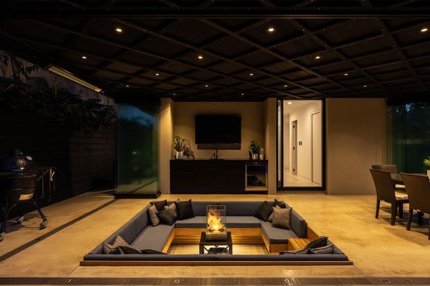 A sophisticated, dimly lit outdoor living and entertainment area with a sunken U-shaped sectional lounge surrounding a central fire feature. The space has a dark coffered ceiling with recessed spotlights, warm beige tiled flooring, dark built-in media cabinetry, large glazed openings, and a dining area at the right. Lush planting and an outdoor grill are visible along the edges.