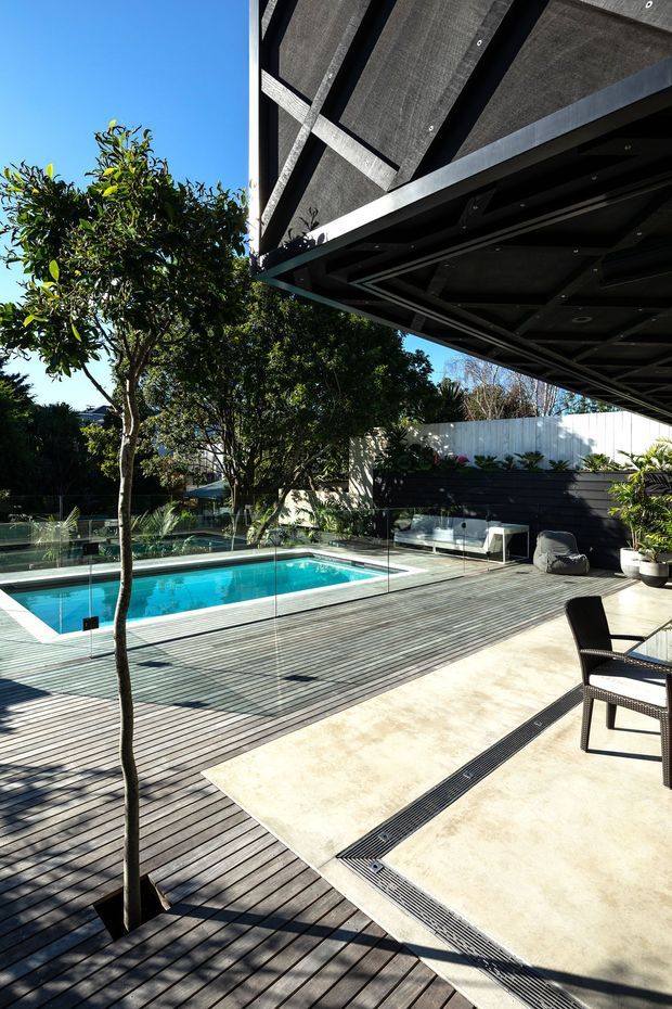 A contemporary outdoor pool terrace viewed from beneath a large covered canopy. The space features a rectangular turquoise swimming pool with frameless glass balustrading, extensive dark timber decking, pale concrete or stone paving, built-in outdoor seating, potted planting, and dark-framed outdoor furniture. Strong sunlight contrasts with the shaded, exposed canopy structure overhead.