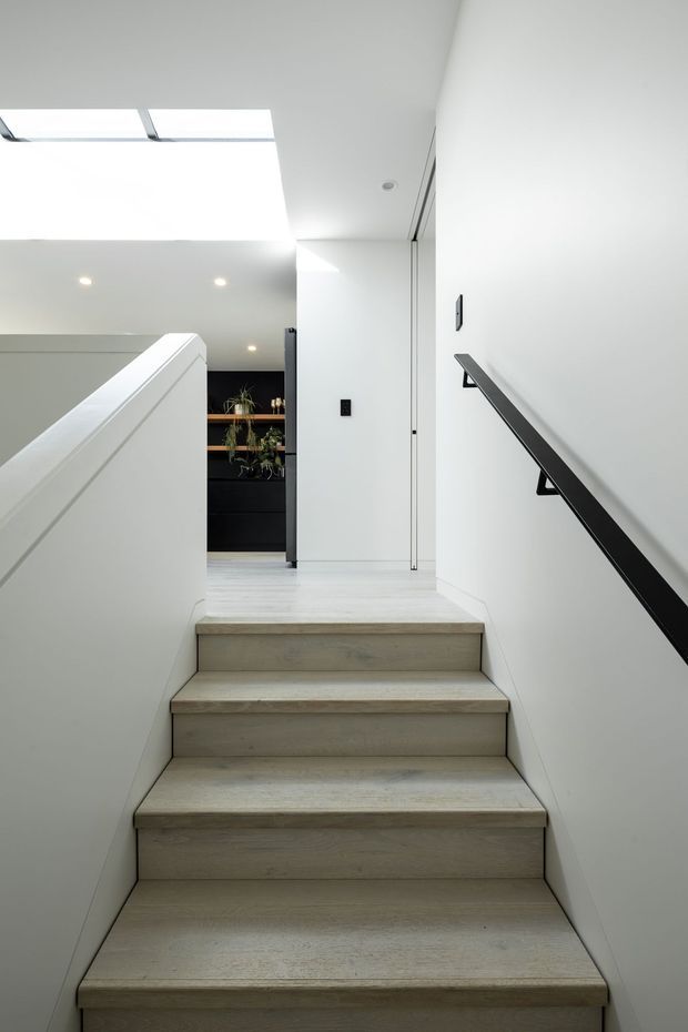 A bright, minimalist stairway leading to an upper landing. The stairs and surrounding walls are finished in pale timber tones and white, with a sleek black handrail on the right wall. A large skylight and recessed ceiling lights provide strong natural and artificial illumination. At the landing, a glimpse of dark built-in cabinetry and indoor plants is visible.