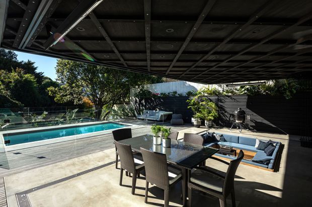 A sophisticated outdoor entertaining area beneath a large dark slatted pergola or roof structure. The space includes a rectangular swimming pool with glass fencing, a dining table with woven chairs, built-in lounge seating with cushions, potted plants, dark feature walls, and concrete and timber-look outdoor surfaces. Strong sunlight creates a high-contrast, resort-like atmosphere.