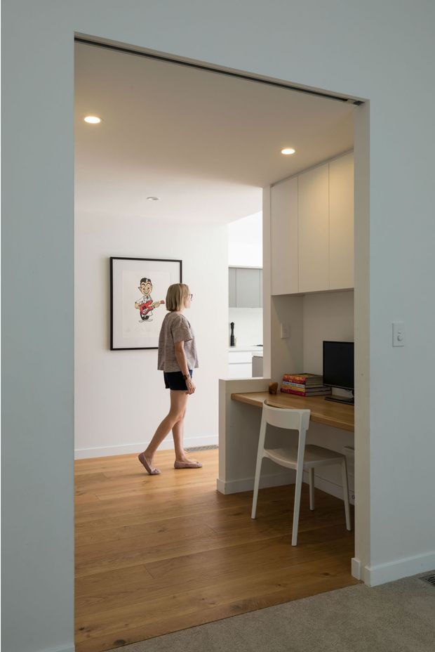 A bright, compact home office nook viewed through a wide doorway, with a built-in timber desk, white chair, overhead cabinetry, computer monitor, and books. The adjoining open area has white walls, warm timber flooring, recessed ceiling lights, and framed artwork. A woman is walking through the space.