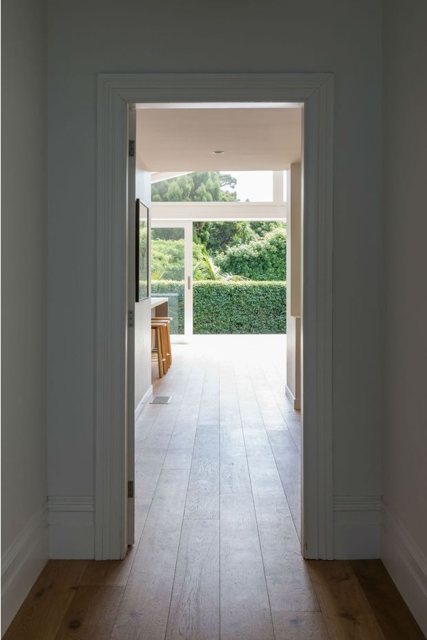 View through a white-trimmed interior doorway into a bright, open-plan living or kitchen area leading to a glazed outdoor space and dense landscaped greenery. The foreground hallway has warm timber flooring, while the adjoining room has pale wide-plank flooring, white walls and ceiling, and a partial timber stool or seating area. The scene is clean, airy, and naturally lit.