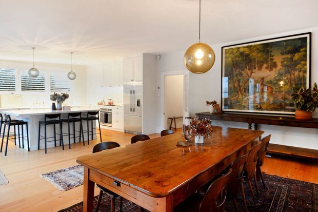 Bright open-plan dining and kitchen interior with a large rustic timber dining table and leather dining chairs in the foreground. The white kitchen includes an island with bar seating, integrated appliances, pendant lighting, and shuttered windows. A large framed artwork, timber console, plants, and layered rugs add warmth to the dining area.