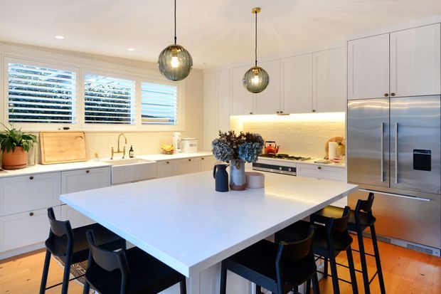 Bright, spacious contemporary kitchen with white shaker-style cabinetry, a large white island with integrated seating, stainless-steel appliances, timber flooring, plantation shutters, and two spherical pendant lights. Black stools and dark hardware provide contrast, while under-cabinet lighting adds warmth.