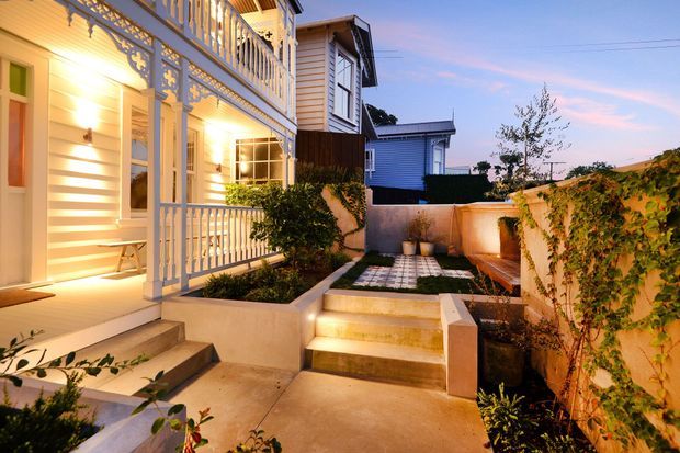 A warmly lit exterior courtyard and entry to a traditional timber-clad home at dusk. The scene features a covered veranda with ornate decorative balustrades, white weatherboard walls, illuminated steps, patterned paving, raised garden beds, and vine-covered boundary walls.