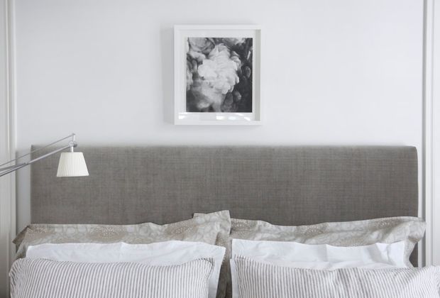 A close-up view of a neatly styled bedroom centered on a tall grey upholstered headboard. Layered white and grey bedding with striped cushions fills the foreground, while a slim adjustable wall lamp and monochrome floral artwork add restrained decorative details against pale walls.