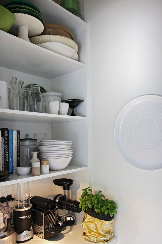 A compact white open-shelving storage area displaying stacked plates, bowls, glassware, cookbooks, and small kitchen appliances. A black coffee machine and blender sit on the lower counter beside a potted herb, while a large decorative white plate is mounted on the adjacent wall. The space is bright, tidy, and styled with predominantly white ceramics and dark appliance accents.