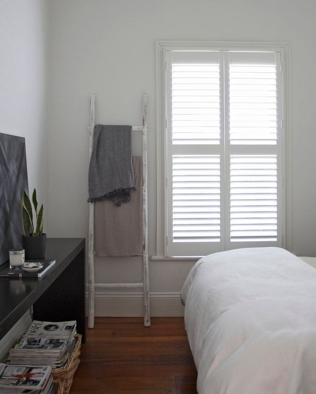 A calm, compact bedroom with white walls, a white bed, dark built-in or low-profile furniture, warm timber flooring, and plantation shutters over a tall window. A rustic ladder displays folded towels or blankets, while a potted snake plant and stacked magazines add understated character.