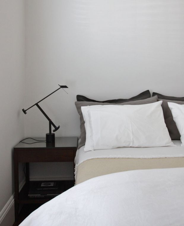 A close-up view of a calm, minimalist bedroom with a neatly made bed in white bedding, dark grey pillows, a dark wood bedside table, and a black adjustable desk lamp. The room has plain light walls, simple trim, and a restrained neutral palette.