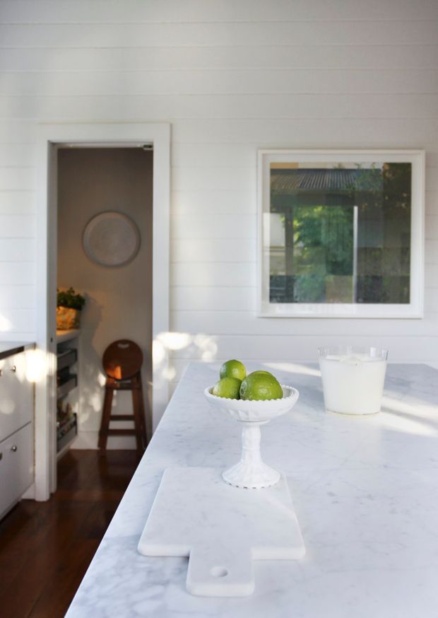 Bright, narrow kitchen interior with white horizontal wall panelling, a large pale marble or stone island benchtop, white trim, and a framed window. A doorway opens to an adjacent room, while a pedestal fruit bowl and glass of milk sit on the island.