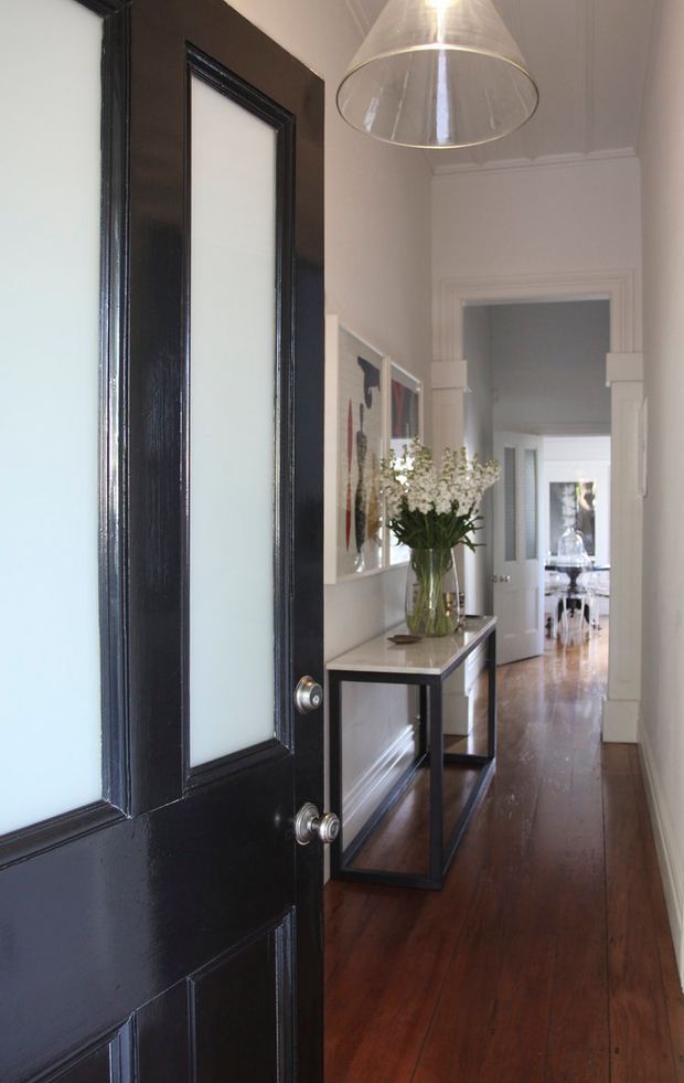 A refined residential hallway viewed through an open black-panelled door. The narrow corridor has polished dark timber flooring, white walls and ornate white architectural mouldings, with a slim console table, framed artwork, a glass vase of flowers and a large translucent pendant light. Additional bright rooms are visible along the hallway.