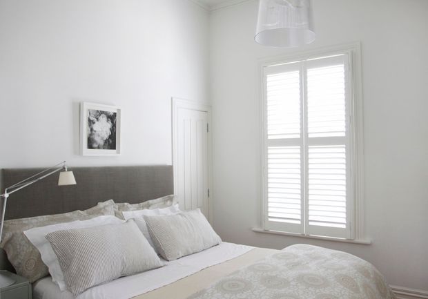 Bright, compact bedroom with a grey upholstered headboard, white bedding, patterned neutral bedspread, striped pillows, a slim wall-mounted reading lamp, white paneled door, framed monochrome artwork, and a large shuttered window. The room has a calm, understated neutral scheme with abundant daylight.