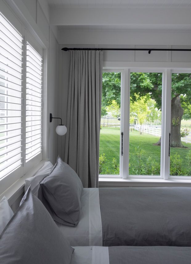 Bright, serene bedroom corner with a neatly made bed in soft grey bedding, a black wall-mounted reading lamp, full-height grey curtains, white plantation shutters, and large white-framed windows overlooking a leafy lawn and fenced garden. The room has white architectural detailing and a calm, restrained palette.