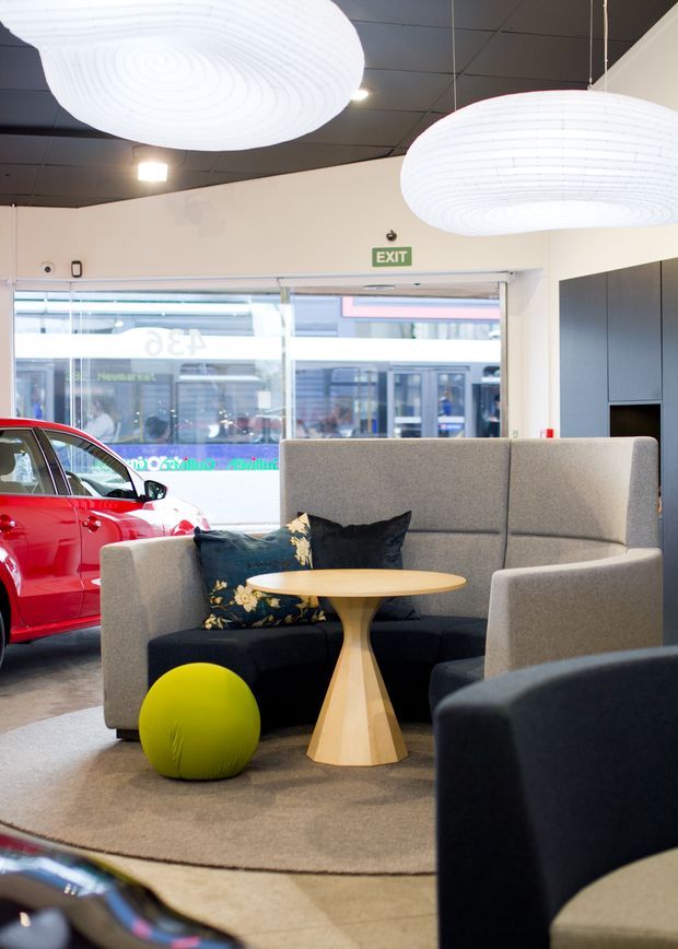 A contemporary commercial showroom or dealership lounge with a grey upholstered high-backed seating booth, black cushions, a light timber pedestal coffee table, and a bright lime-green ottoman on a large neutral rug. Oversized white pendant lights hang from a dark tiled ceiling. A red car is displayed beside the seating area, with full-height glazing and an EXIT sign visible in the background.