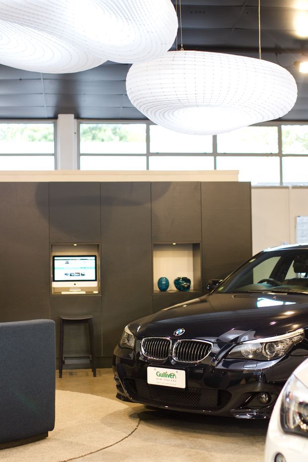 Interior of a contemporary automotive showroom featuring a dark BMW vehicle in the foreground, dark built-in display cabinetry with recessed niches and a wall-mounted screen, large industrial-style windows, and oversized white pendant lights.