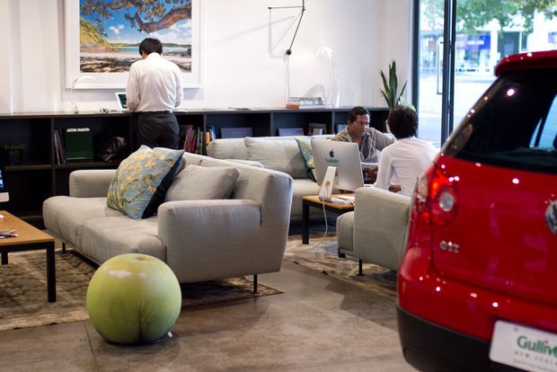 A contemporary showroom or studio lounge with multiple upholstered sofas and armchairs arranged around low tables, a long dark built-in shelving unit, artwork, and a wall-mounted articulated lamp. Several people are using laptops in the seating area. A red compact car is prominently displayed inside near full-height glazing, suggesting an automotive or lifestyle commercial space. The room has polished concrete flooring and a neutral, softly lit interior.