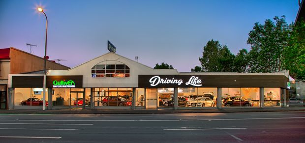 Street-facing commercial showroom with a long, low facade, illuminated signage, extensive storefront glazing, and vehicles displayed inside. The building combines dark horizontal cladding with a light rendered central gabled section and is photographed at dusk from across a multi-lane road.