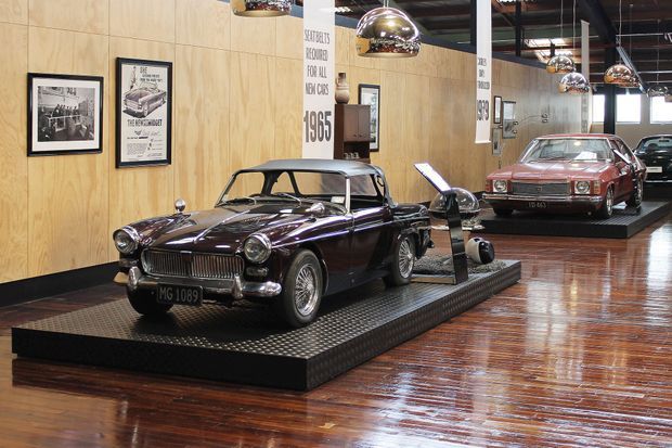 Interior of a polished automotive showroom or museum displaying classic cars on raised black platforms. The space features warm plywood wall panelling, glossy dark timber flooring, framed automotive posters, exposed dark ceiling services, and multiple chrome pendant lights. Additional vehicles and their reflections are visible in the background and mirrors.