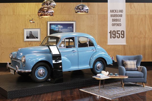 A retro-styled commercial display space featuring a light-blue vintage car mounted on a raised black platform. The room has warm plywood wall panelling, polished wood flooring, framed photographs, chrome pendant lights, and a small lounge arrangement with a blue armchair, coffee table, and rug. A wall banner references the Auckland Harbour Bridge opening in 1959.