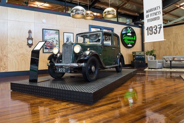 A polished commercial interior styled as a vintage automotive showroom or service-themed hospitality space. A restored black vintage car sits on a raised diamond-plate metal display platform, surrounded by plywood wall panelling, framed automotive artwork, retro signage, pendant lights, exposed ceiling services, and polished timber flooring. A small lounge area is visible at the rear.